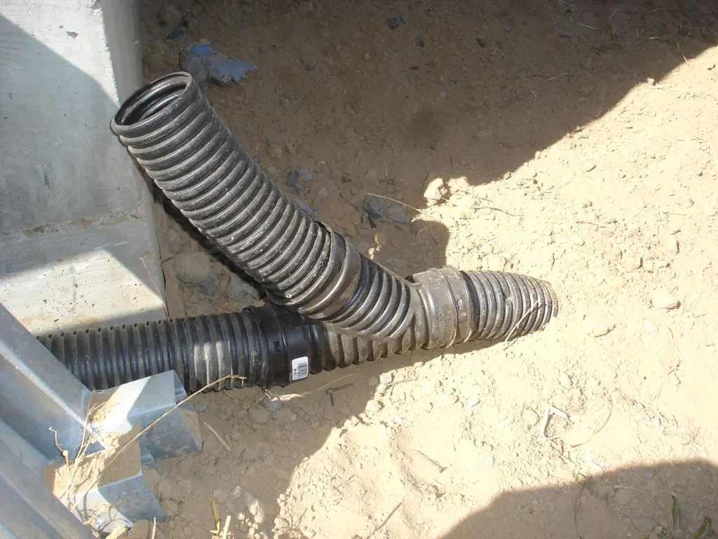 Blackland clay drainage beside a foundation: corrugated drain pipes joined at a fitting next to a house wall